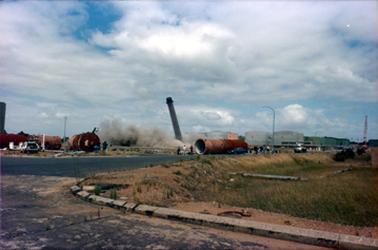 This image shows an industrial site with a tall chimney leaning to the left.  A large area is covered with thick smoke.  Large grey buildings are in the background.  Pipes and tanks are on either side of the tower.  A street is in the foreground.  Pipes are lying in the grass.  People can be seen standing on the road watching the demolition.  