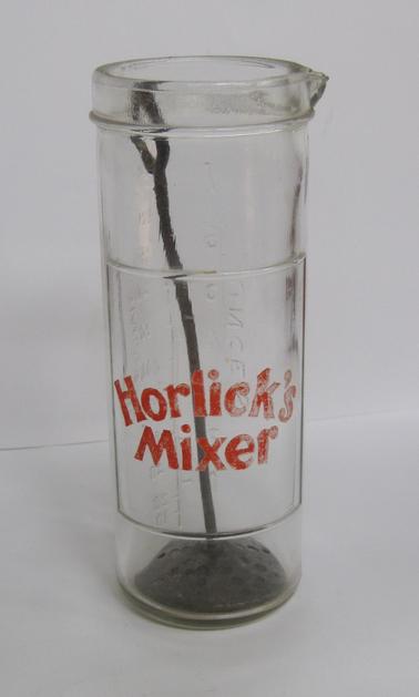 Cylindrical glass containerwith lip at top.  No lid.  Measurements in ounces and grammes.  Impressed in glass in red lettering:  “Horlick’s Mixer”.  Metal plunger with long handle attached to circular disk with holes to mix the Horlick’s powder (Horlick’s brand of malt) and milk.