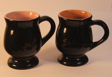 Two ceramic mugs with curved bulbous body on a short rounded base, with a rounded handle on one side. The mugs have been glazed with a black exterior. Object 1.1 has a brown interior with the word “Dad” on the outside. Object 1.2 has a pink interior with the word “Mum” on the outside.
