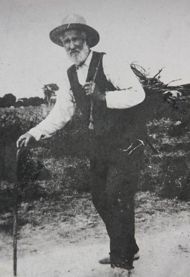 Black and while photograph of Robert Rowley with walking stick, carrying a bundle of firewood on his back.  