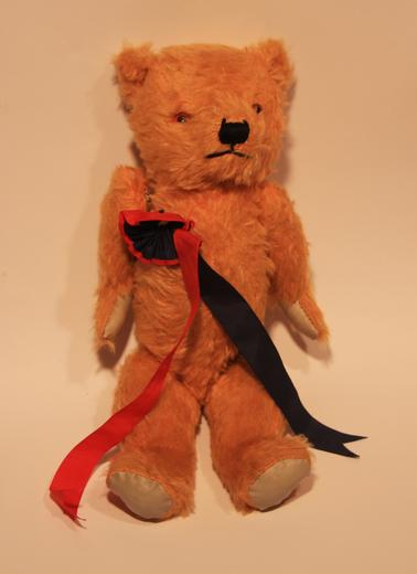 Common teddy bear with artificial fur.  Yellow.  Button eyes, embroidered mouth and nose.  Rosette and ribbons are Rye School colours for sports day.