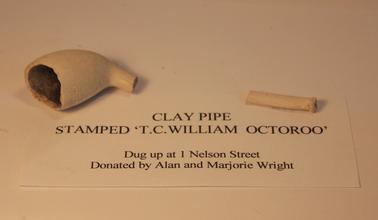 A broken white clay pipe. The bowl is oval in shape and the stem is narrow and cylindrical. The centre piece of the stem is broken and missing.  Inscriptions on the card with the pipe reads: “T C William, Octoroo”.