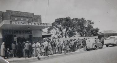 Queue outside Rowley’s butcher shop on Pt Nepean Road on 4 January 1949.  Rye Store and chemist sign on right.  Austin car and utility parked on road. 