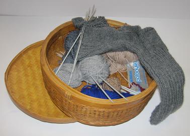 Round basket, with lid, containing selection of knitting wool and needles.  Unfinished man’s sock on four needles.