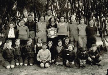 Head Teacher Lionel Martin with pupils (in two rows). One of the pupils is holding the Sports shield 1932. The group is standing in the Rye School grounds.  
Inscription on back reads: “Rye State School.  Mr Lionel Martin with pupils.”