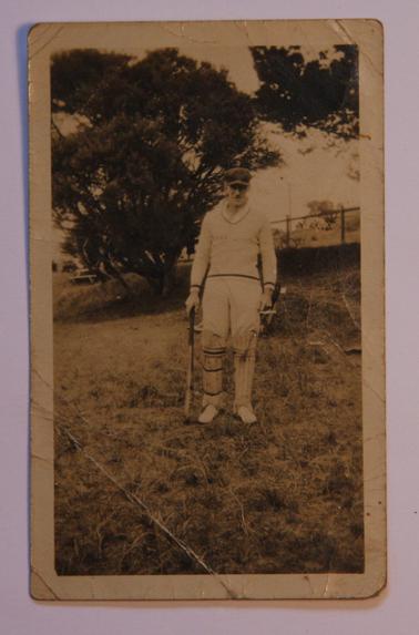 Alan Sullivan in cricket whites: cap, pads, with bat, at cricket ground.