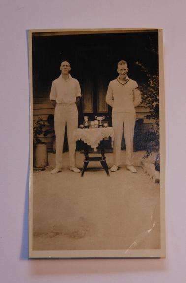 Jack and Alan Sullivan in cricket whites, standing each side of a small table with crocheted cloth.  There are 6 trophies for cricket on the table.  (They are Jack’s)