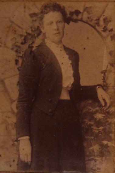 Nellie Sullivan in dark skirt and open fitted jacket; white blouse with ruffle.  Standing in front of an arch within a wall.