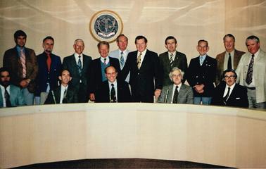 Photograph Shows the eleven Councillors and Council Officers of Flinders Shire ca.1980.  Council emblem on wall at back.