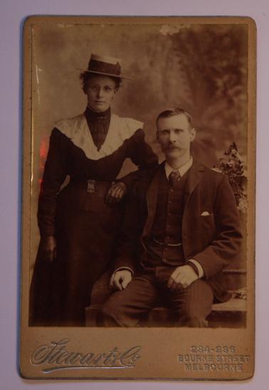 Sepia photograph of Nellie and Jim Sullivan.  Woman standing, wearing a black dress, white shawl, kid gloves, boater hat (straw) with black ribbon.  Man - seated, wearing suit, shirt and tie and waistcoat with watch chain.