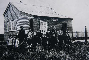Photo of 12 children (student at left back row : Eileen Hill), plus male teacher outside Boneo School (No. 1184). Date unknown.