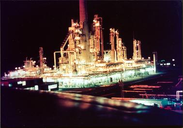 This is a night time photo of an oil refinery.  The entire complex, which includes many tall towers, is fully lit and the surrounds are black.  