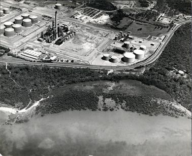 This black and white aerial photo shows an oil refinery in the top half and a bay and wetlands in the foreground.  A curved road separates the two at the centre of the image.  