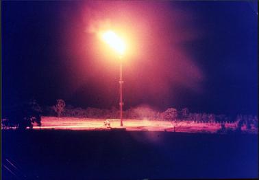 This is a night time photo showing a very tall flare stack at the centre of the image.  Flames are billowing from the top lighting up the ground underneath.  A line of trees are in the background and a fence is on the far right-hand side of the image.  The surrounds are pitch black.  