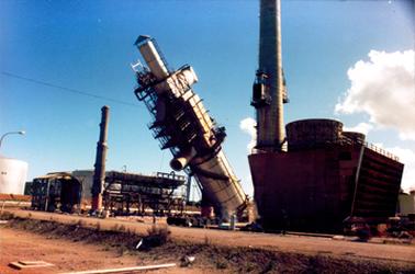 This image shows a large tower leaning over to the left.  A tall chimney stack is to the left and part of another large tower is to the right.  A large building is on the far right and a white tank is on the far left-hand side.  A road is in the foreground.  