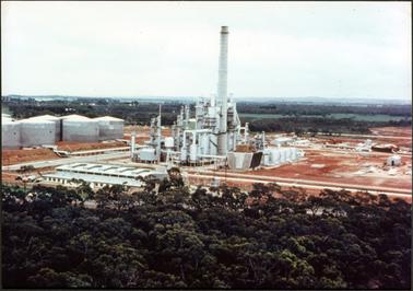 This photo shows an oil refinery with a very tall stack, at the centre of the image surrounded by tanks and buildings.  Heavily treed areas are in the foreground and behind the refinery.  Water and land is in the far background.  
