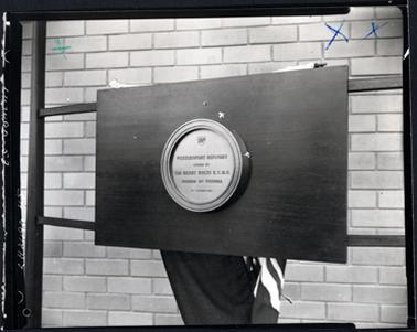 This image shows a circular plaque at the centre of a board which is attached to a frame.  It stands in front of a brick wall.  Part of a flag can be seen is hanging behind the board.  The following is printed on the plaque: “BP Westernport Refinery opened by Sir Henry Bolte K.C.M.G. Premier of Victoria 21st October 1966”. Two blue biro X ARE IN THE TOP RIGHT-HAND CORNER.  A GREEN X IS IN THE TOP LEFT-HAND CORNER.  The left-hand edge of the photograph has been roughly cut.