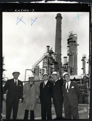 This image shows five men standing in front of a refinery, smiling and looking up.  They are all wearing suits and hard hats.  The man second from the left is wearing a rain coat and the man second from the right, is pointing upward.  Across the top of the image are three X written in blue biro.  Printed in white on the black border at the top is: “LH2490-3”. The edges of the photograph have been roughly cut.