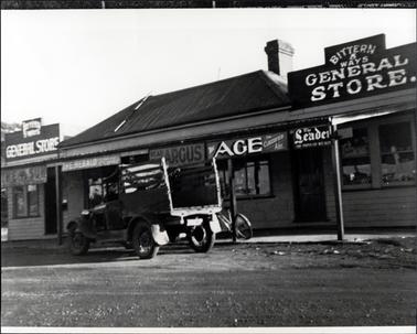 This image shows a shop front covered in signage.  The section in the middle has a brick frontage and the two sections on either side are timber.  Windows and doors are across the front.  Four verandah posts are along the footpath and an old delivery truck is parked beside the shop.  A bicycle is leaning against a post beside the truck.  Signs across the front of the shop read as follows: “Bittern 5 Ways General Store, The Sun, The Herald, Globe, Read Argus, The Age, Classified Ads, and The Leader The Popular Weekly”.  A gravel street is in the foreground.  