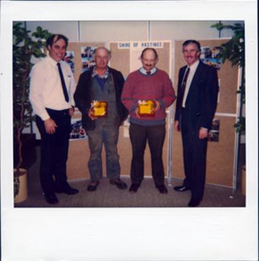 This image shows four men standing in a row in front of a screen, smiling for the camera.  The two men in the centre are holding gift boxes.  The man on the left is wearing a long sleeved white shirt and tie and the man on right is wearing white shirt tie and suit.  A sign which reads: “Shire of Hastings” is at the top of the screen behind the group.  Potted plants are on either side.   