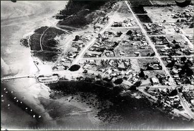 This image shows an aerial photograph of a foreshore, jetty and boat harbour to the left and a town to the right.  Boats dot the water in the foreground. 
