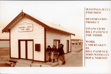 This image shows three men leaning on a railing beside a small shed.  They are all smiling for the camera.  The man on the right has his left hand on the top of his hat.  The board on the front of the shed reads: “Circa 1866 Hastings Jetty Fish Shed Restoration 1989”. Part of a jetty, water and tanks can be seen in the background.  The following is printed on the right-hand side of the photograph: “Hastings Jetty Fish Shed Restoration Project Financed by Bill Patience The Third.  Work undertaken by Bill Patience John Doug Norton’.