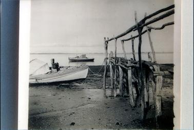 This image shows a wooden boat on the sand, tied to a roughly constructed elevated walkway. Another similar wooden boat is in the water.  Land can be seen in the distance.  