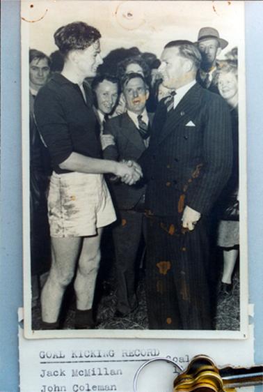 This image shows two men shaking hands, surrounded by a large group of men and women.  The man on the right is wearing a pin-striped double-breasted suit.  The man on the left is wearing a dark coloured football shirt and dirty white shorts.  Typed on a piece of white paper at the bottom of the photograph is: “Goal kicking record Jack McMillan John Coleman Goal”.  Part of a set of keys are also part of the image. 