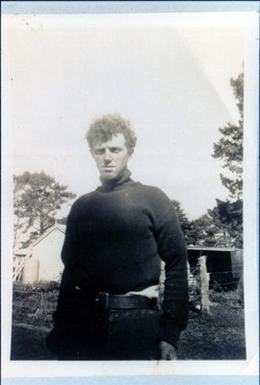 This image shows a young man standing with his arms to his sides and a serious look on his face.  He has a mop of curly hair.  He is wearing a dark coloured high-necked pullover, dark coloured trousers and a wide leather belt.  Part of a weatherboard building and a shed can be seen behind the barbed wire fence.  Tall trees are on either side of the image.  