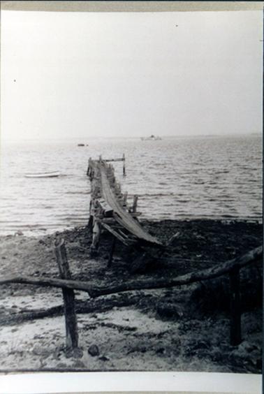 This image shows an old wooden jetty.  The photo is taken from the shoreline looking down the middle of the jetty.  Part of a fence made from branches is in the foreground.  A small boat is to the left of the jetty and a large boat is in the distance.  