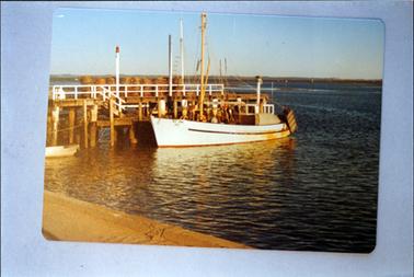 This image shows a fishing boat beside a wooden jetty. Two rows of fishing pots are on the jetty.  Land can be seen in the distance and a concrete barrier in the foreground.    
