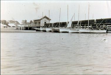 This image shows a line of seven wooden fishing boats beside a wooden jetty.  The photo is taken from the water looking back to land.  A small shed is on the jetty and other buildings can be seen on the shore.  Fishing nets are hanging over the top rail of the jetty.