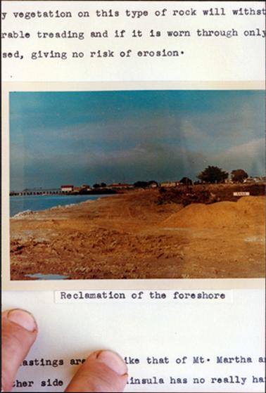 This image shows a photo at the centre with printing top and bottom, including the ends of two fingers.  The photo shows land works in the foreground, water, a jetty and shed to the left and buildings and trees to the right. The caption under the photo reads: “Reclamation of the foreshore”.