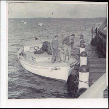 This image shows two men standing on a boat, looking down into the water.  The boat is tied up to a large white post beside a jetty.  Both men are wearing jumpers.  The man on the left is wearing long pants and the other man is wearing shorts.  Part of the jetty can be seen on the right-hand side of the photograph and four boats can be seen in the distance on the left.  Land can be seen in the far distance.  