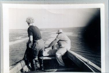 This image shows part of a wooden fishing boat, with the back view of a woman and a man working with fishing nets.  The woman has fair curly hair, a short sleeved dark coloured shirt, light coloured pants and high waders.  The man is wearing a hat and light coloured long sleeved jacket and pants.  