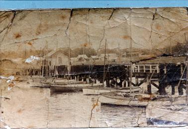 This cracked and faded image shows a jetty with fishing boats lined up along the side, looking back to the town.  Fishing nets are hung over the rail of the jetty.  A small shed is on the jetty and buildings can be seen on the land.  