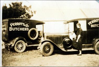 This image shows two vans parked facing each other.  They have ‘Perrott & Co Butchers’ painted in white on their vans.  Both vehicles have running boards and no doors.  The van on the left has wooden spokes on the wheels.  Both vans have spare tyres attached to their sides.  A woman, dressed in black, is standing beside the van on the right.  She has one foot on the running board.  