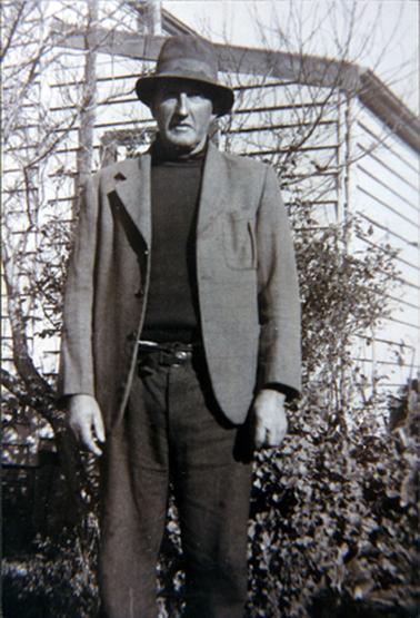 This image shows a man standing in front of a timber house, posing for a photo.  He is wearing dark coloured trousers and pullover with a lighter coloured sports coat.  His hat is shading his eyes and he has a serious look on his face.  