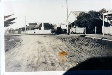 This street scene shows an wide dirt street in the foreground and a line of houses and shops on the right-hand side.  A motor vehicle is parked on the footpath and a black dog is sitting beside a white picket fence.  Three tall power poles are along the side of the street.  