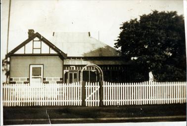 This low-set house has a steep sloping iron roof with a closed-in verandah on right-hand side and a high gable on the left.  A brick chimney can be seen behind the gable.  A white picket fence runs along the front of the property and a trellis is behind the small front gate.  A large tree is on the far right-hand side and a footpath is in the foreground.  