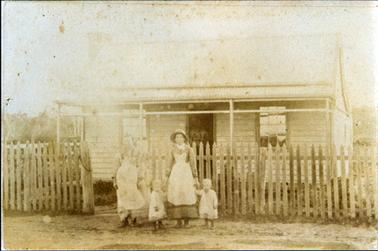 This faded image shows two people and two small children, standing in front of a wooden picket fence on the footpath, posing for a photo.  A small timber cottage with a four posted verandah is behind the fence.  The children are exactly the same size and look like twins.   