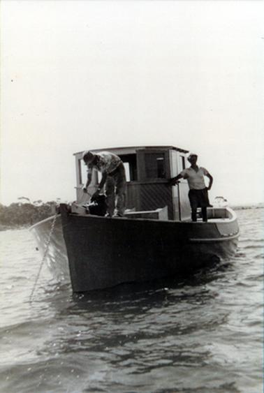 This image shows a boat at anchor with two men on board.  The man on the right is leaning on the high wooden cabin and the other man is bending over at the front of the boat.  The shoreline can be seen on the left-hand side.  
