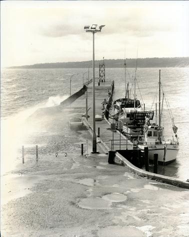 This sepia portrait photograph shows three fishing boats tied to a wooden jetty.  The sea is choppy and waves are breaking over the left-hand side of the jetty.  A motor vehicle is standing beside the jetty and a small up-turned boat is lying on the jetty.  A very tall pole holding three lights is at the start of the jetty.  A sign on the jetty reads: “Permit vehicles only beyond this point”.  Land is in the background and deep potholes are in the foreground.  The boat in the foreground is named “TV1HELLAS”.  