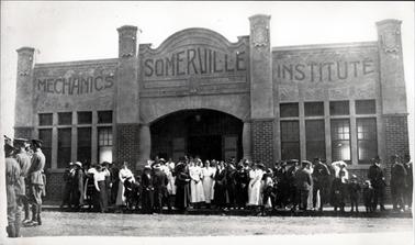 Approximately fifty people are standing in front of the Somerville Mechanics Institute. The crowd consists of men, women, children, soldiers and nurses.  The building has an imposing front consisting of four very tall pillars.  The bottom half is brick and the top section is concrete.  Large sets of windows are on either side of the imposing entry and front doors.  