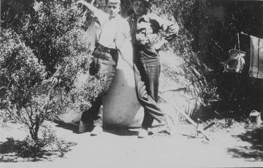 Merricks Foreshore. Two young men leaning against each other in front of a tent. L.H. figure has one arm raised. Bare-headed, long trousers with braces, shirt sleeves rolled up. R.H. figure has one hand on hip, wearing hat, glasses, long sleeved shirt, long trousers with braces. Low bush L. foregrouno date. Towel and carry can hanging on rope strung across trees to right. Container on grouno date.