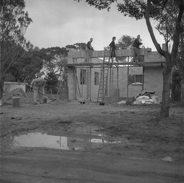A building under construction: on scaffolding at ceiling height.two men and one woman laying cement blocks while one man works on the grouno date.  A tree and a water puddle in the foreground and trees in the backgrouno date.
