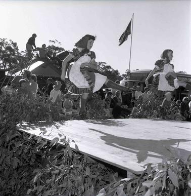 Two girls on an outdoor stage festooned with gum leaves performing Irish Dancing.  People in the background,