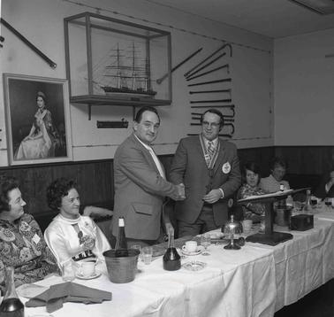 Two standing men shaking hands at a function dining table with others seated.