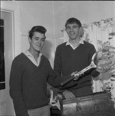Two young menwearing open necked shirts and dark jumpers, one with a tennis racket.