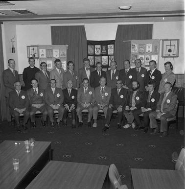 Twenty two men in suits posing for an indoor photograph in front of a display of club banners.